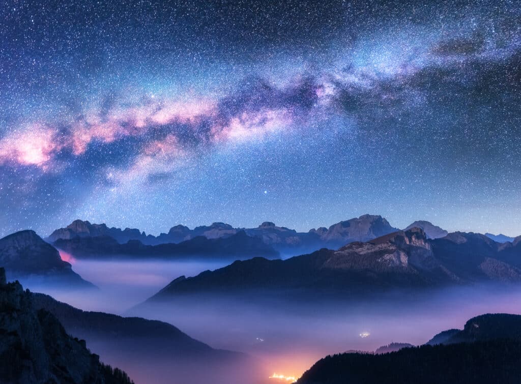 The cosmos, Milky Way above mountains in fog with city illumination, a purple night sky. Used to show life, nature, and the universe are one, by Joanne Z. Tan.